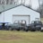 A collection of vintage car and truck bodies is scattered in front of a large white garage with a green metal roof. The scene is set on a grassy field with leafless trees in the background, indicating a late autumn or winter season. Various car parts, including an old blue car body and a gray truck, are visible. The vehicles appear to be in various stages of disrepair or restoration.